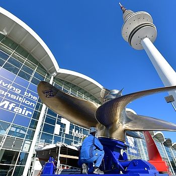 Ein goldenes Windrad, welches vor dem Hamburg Messe Gebäude installiert wird. Das Messegebäude wird schräg von unten gezeigt. 