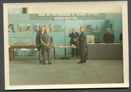 Historical picture of the information stand of the Association of Ship Engineers in Hamburg E.V.. With two tables and several pictures hanging on the wall.