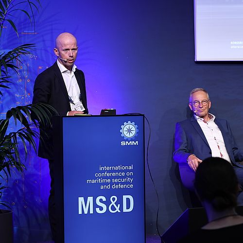 Frontalaufnahme eines Speakers am Pult und eines weiteren Speakers, der neben ihm auf einem Stuhl sitzt, auf der MS&D-Konferenz der SMM 2024.