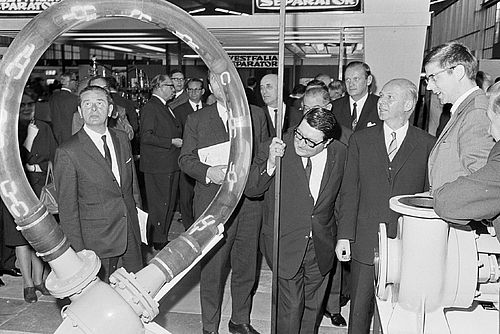 A black-and-white photograph of a trade fair from an earlier period, taken from the audience perspective, with everyone looking at an exhibition piece