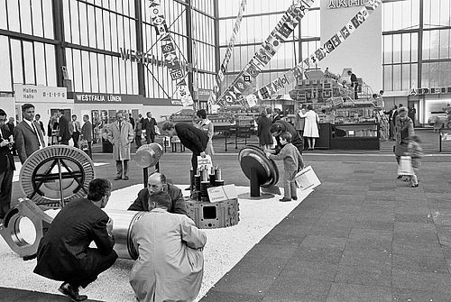 A historical picture of a trade fair in Westfalia Lünen. There are several large parts of a ship on display with many visitors to the fair, both adults and children.