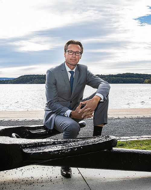 A man in a gray suit sits on a large anchor. Behind him is a landscape with a walkway, water, a blue cloudy sky and green mainland.