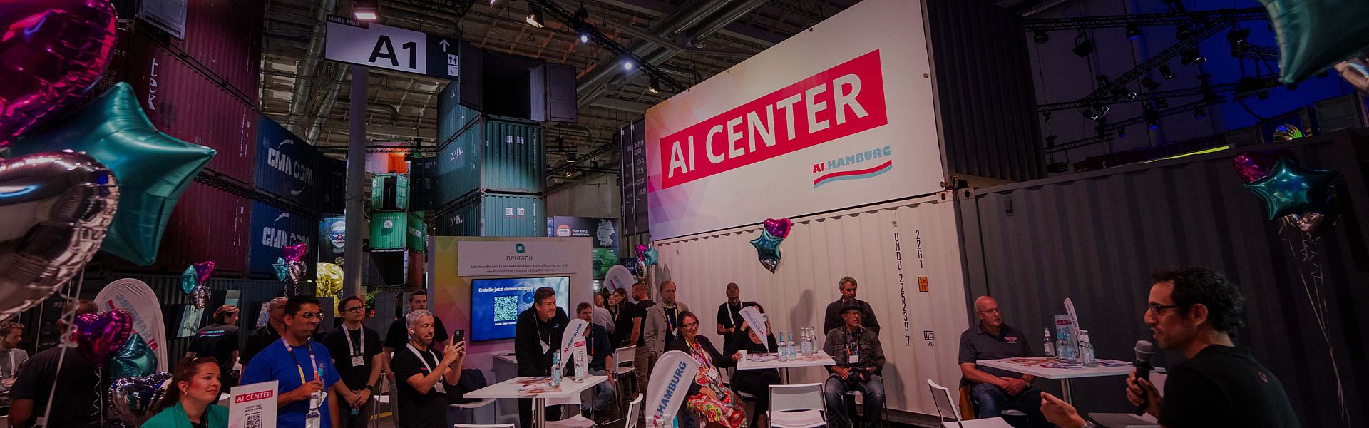 A side-view image in the AI Center Area with someone speaking into a microphone. Audience members sit at tables while one person films