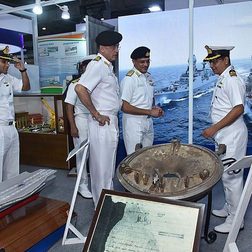 [Translate to EN:] Das Bild zeigt eine Gruppe von Marineoffizieren in Uniform, die sich auf einer Ausstellung über maritime Technologie unterhalten. Im Hintergrund ist ein großes Bild von Kriegsschiffen auf See zu sehen, und es werden Modelle von Schiffen sowie historische maritime Artefakte ausgestellt.