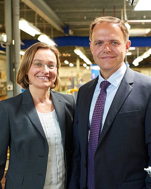 Tanja and Ronald Hoppmann look into the camera smiling and wear gray suit jackets. In the background you can see a kind of warehouse..